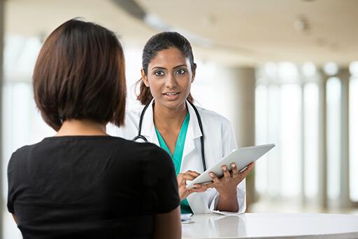 Une femme avec un tee-shirt noir et avec des cheveux courts et noir est face à un docteur. Le docteur tient une tablette