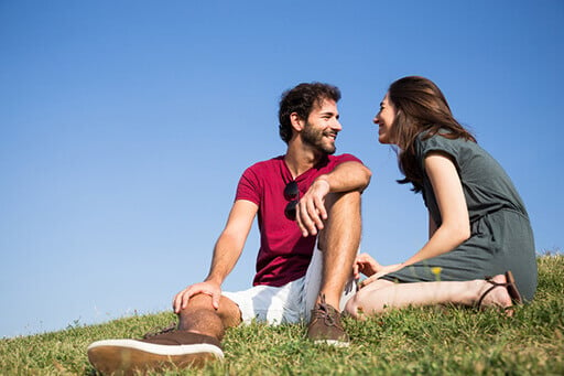 Ciel bleu en fond. Une femme et un homme se sourient, ils sont assis sur de l'herbe